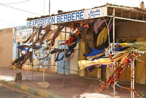 Am Rande der Sahara wirbelt der Wüstenwind einen Souvenirladen auf, den Imazighen am Rande einer Straße errichtet haben. Die Stadt M