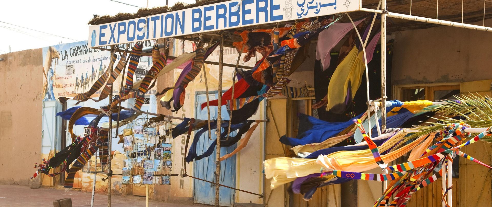 Am Rande der Sahara wirbelt der Wüstenwind einen Souvenirladen auf, den Imazighen am Rande einer Straße errichtet haben. Die Stadt M